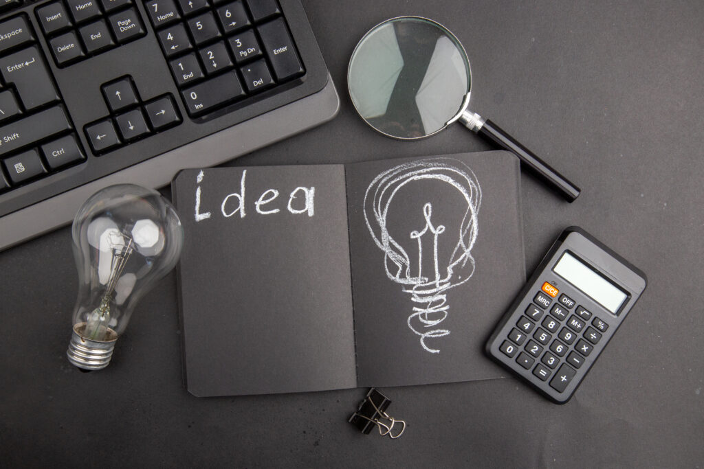 top-view-idea-written-black-notepad-keyboard-lupa-binder-clip-light-bulb-calculator-dark